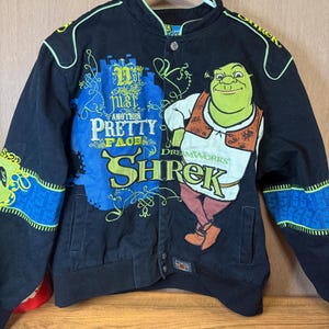 Pode incluir: Casaco preto do Shrek com fecho de botões. O casaco apresenta um grande gráfico do Shrek, o texto "Another Pretty Face" e o logótipo da DreamWorks. As mangas têm detalhes azuis e amarelos, e o casaco está pendurado num cabide branco.