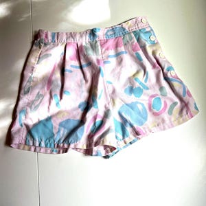 May include: Pink shorts with a colorful abstract pattern in blue, pink, and yellow. The shorts have a button closure and side pockets. The fabric appears to be lightweight.