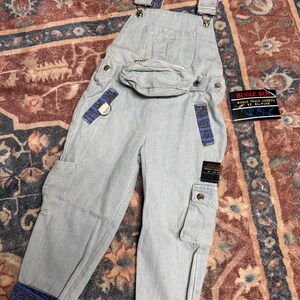 Op de afbeelding: Lichtblauwe spijker tuinbroek met een ingebouwd heuptasje, zijzakken en blauwe stoffen accenten met patronen op de schouderbanden en boorden. Een zwart label vermeldt BUGLE BOY WORLD PEACE COUNCIL N° SP RL-9130, passend bij het label op de broek.