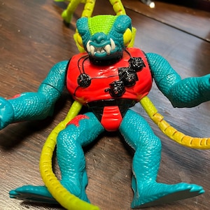 May include: A vintage action figure of a reptilian alien creature. The figure has a teal body, a red torso, and yellow appendages. The head is green and blue with sharp white teeth. Black details are on the chest.