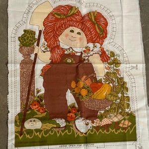 Könnte beinhalten: Vintage Stoffpaneel mit einer Cabbage Patch Kid-Illustration. Das Design zeigt eine rothaarige Puppe in braunen Latzhosen, die eine Schaufel und einen Korb mit Produkten hält. Der Text "LEAVE OPEN FOR STUFFING" ist sichtbar.