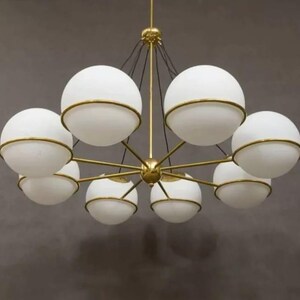 Pu&ograve; includere: Un lampadario moderno con otto paralumi sferici in vetro bianco, ciascuno circondato da una fascia metallica color oro. L'apparecchio ha un mozzo centrale color oro con bracci radiali, sospeso dal soffitto.