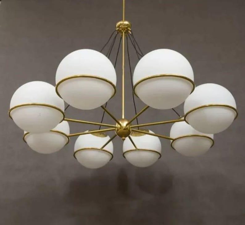 Pu&ograve; includere: Un lampadario moderno con otto paralumi sferici in vetro bianco, ciascuno circondato da una fascia metallica color oro. L'apparecchio ha un mozzo centrale color oro con bracci radiali, sospeso dal soffitto.