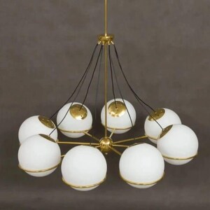 Pu&ograve; includere: Un lampadario moderno con otto paralumi a globo bianchi, ciascuno circondato da una fascia color oro. L'apparecchio ha un mozzo centrale e bracci di supporto color oro, con cavi neri che sospendono i paralumi. Il design &egrave; elegante e contemporaneo.