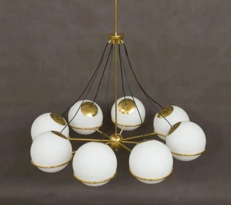 Pu&ograve; includere: Un lampadario moderno con otto paralumi a globo bianchi, ciascuno circondato da una fascia color oro. L'apparecchio ha un mozzo centrale e bracci di supporto color oro, con cavi neri che sospendono i paralumi. Il design &egrave; elegante e contemporaneo.