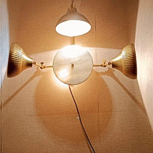 Pode incluir: Uma arandela de parede dourada com três luzes. A luz central tem um abajur de mármore branco e as duas luzes laterais têm abajures dourados. A arandela está montada em uma parede.