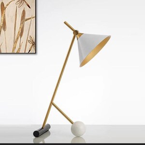 Pode incluir: Uma moderna luminária de mesa com uma cúpula cónica branca e interior dourado. A luminária apresenta uma estrutura angular dourada e uma base esférica de mármore. A base também tem uma peça retangular preta. Uma luminária elegante.