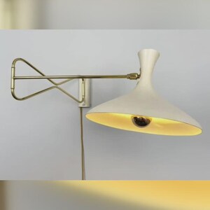 Pode incluir: Arandela de parede creme com braço de latão e abajur flare. A lâmpada tem um design moderno de meados do século, com uma estética simples e elegante. A luminária é montada em uma parede branca.