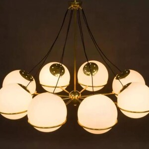 Pu&ograve; includere: Un lampadario moderno con otto paralumi sferici in vetro smerigliato. Ogni globo &egrave; circondato da una fascia metallica color oro e sospeso a un apparecchio centrale con cavi neri. L'apparecchio ha una finitura dorata.