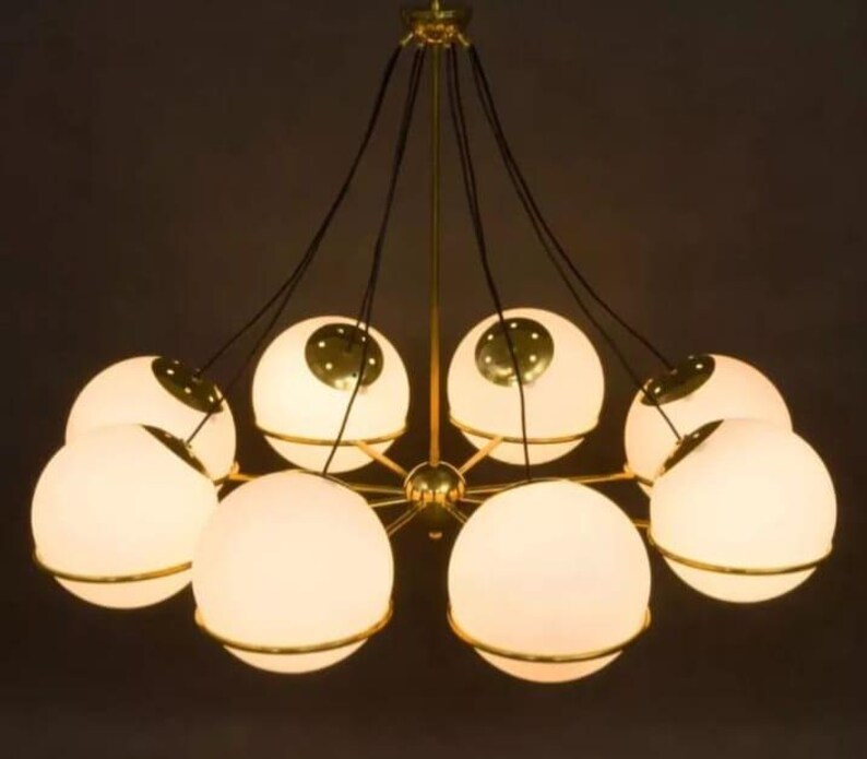 Pu&ograve; includere: Un lampadario moderno con otto paralumi sferici in vetro smerigliato. Ogni globo &egrave; circondato da una fascia metallica color oro e sospeso a un apparecchio centrale con cavi neri. L'apparecchio ha una finitura dorata.