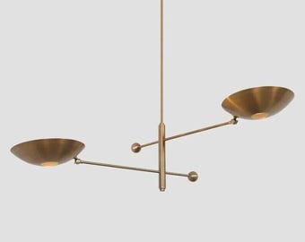 May include: A brass pendant light fixture with two adjustable, angled shades. The fixture has a minimalist design with a central rod and two arms extending out to the shades.