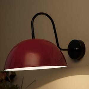 Pode incluir: Arandela de parede vermelha em forma de cúpula com um braço curvo preto e uma base circular. A arandela tem um acabamento brilhante e um interior branco, proporcionando luz para baixo. Montada numa parede de cor neutra.