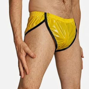 May include: Bright yellow athletic-style shorts featuring a glossy, vinyl-like finish. The garment has a high-cut leg design with contrasting black trim along the edges and a comfortable elastic waistband, providing a sleek and fitted appearance.