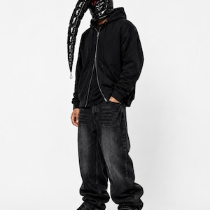 Puede incluir: Una sudadera negra con cremallera, vaqueros de mezclilla gris carbón de corte holgado y zapatillas negras. La cabeza está cubierta por una máscara acolchada negra brillante con una extensión larga y curva en forma de hocico y ojos rojos.