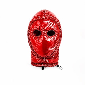Könnte beinhalten: Eine leuchtend rote Sturmhaube aus glänzendem, gestepptem Material im Daunenjacken-Stil. Die Kopfbedeckung verfügt über zwei kreisförmige Augenöffnungen mit dunkler Umrandung und einen Kordelzug am unteren Rand mit einem schwarzen Kunststoff-Stopper.