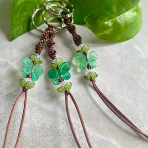 May include: Three green butterfly charm keychains with brown cords and silver key rings. The charms are made of glass and have a white center.