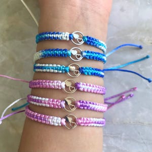 May include: A set of six friendship bracelets with a silver heart charm. The bracelets are made with blue, white, and pink ombre colored cords.