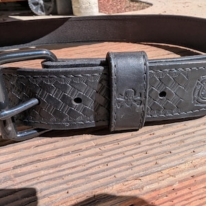 May include: A black leather belt featuring a basketweave pattern, a black metal buckle, and a belt loop embossed with a skull and crossbones design. The belt also displays a stamped emblem of a Maltese cross, commonly associated with firefighting.