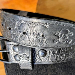 May include: A black leather belt featuring an embossed pattern of multiple skulls and crossbones. The belt includes a dark metal buckle and a matching leather keeper loop, also embossed with a small skull and crossbones design.