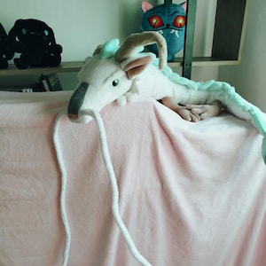 May include: A plush white dragon with long white whiskers, brown horns, and a gray snout rests on a pink blanket. The dragon has a light green mane and a long, segmented body. Other plush toys are visible on shelves in the background.
