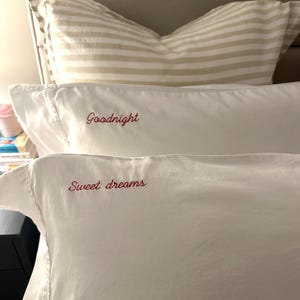 Op de afbeelding: Witte kussenslopen met rode geborduurde tekst. E&eacute;n kussensloop heeft de tekst "Goodnight" en de andere "Sweet dreams". Op de achtergrond is een gestreept kussen te zien.
