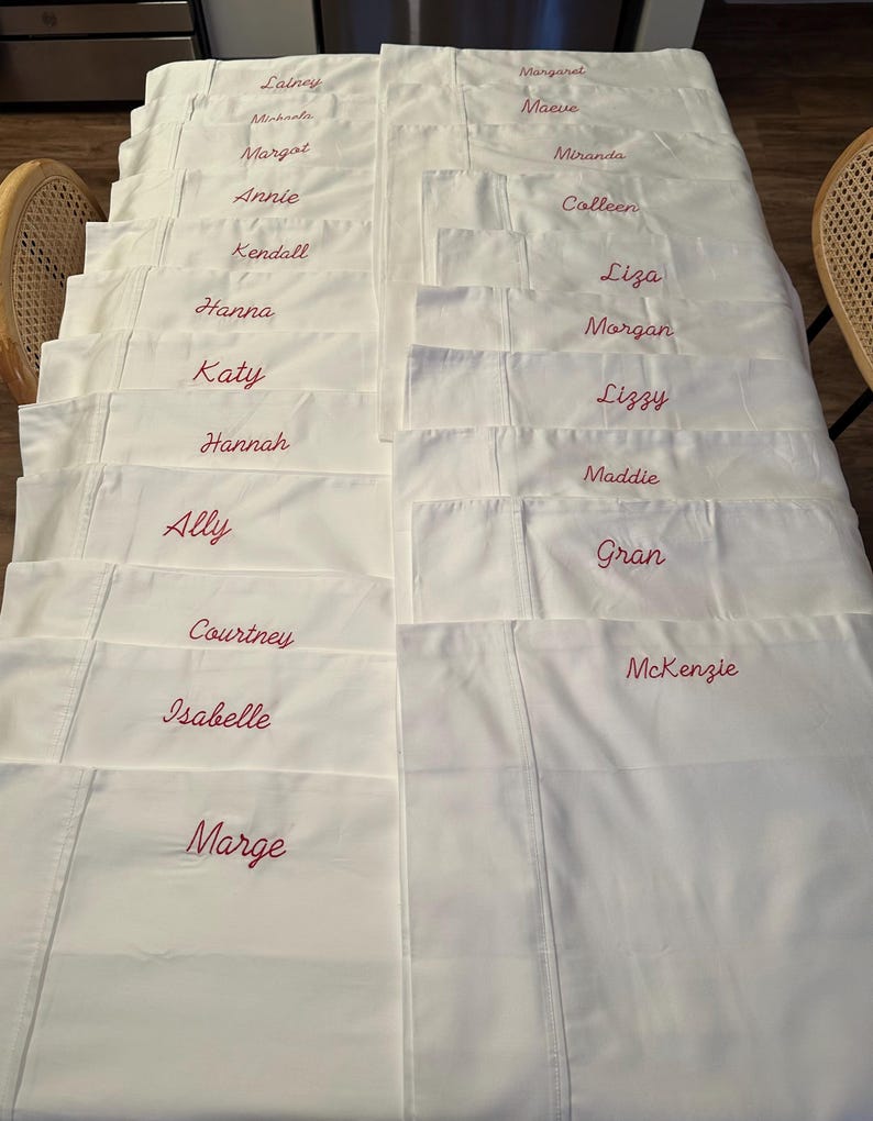Op de afbeelding: Een verzameling witte lakens, elk geborduurd met een andere naam in een bordeauxrode, cursieve letter. De lakens zijn netjes opgevouwen en gerangschikt, wat gepersonaliseerd beddengoed laat zien.