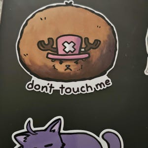 May include: A brown cartoon character with antlers wearing a pink hat with a white X on it. The character has a grumpy expression and the text "don't touch me" is written below it.