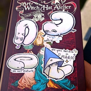 May include: A selection of quirky stickers from "The Art of Witch Hat Atelier" are placed on a book cover. The stickers showcase unique characters and phrases such as "do not disturb" and "do you believe in magic". The book's cover is a deep burgundy hue.