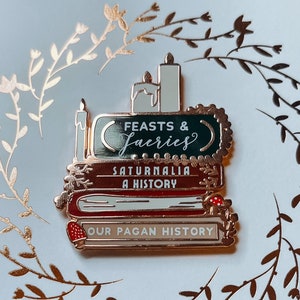 May include: A pin featuring a stack of books with the titles "Feasts & Faeries", "Saturnalia A History", and "Our Pagan History". The pin has a gold border and is set against a light blue background with the text "Belle's Books & Candles" and "the magic of reading".