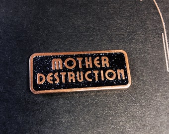 May include: A rectangular pin with the words "MOTHER DESTRUCTION" in copper-colored lettering. The background is black with glitter. The pin has a copper-colored border and is set against a dark background.