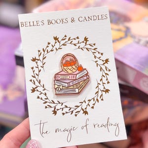 May include: A pin featuring a stack of books with the titles "Little Women", "Emma", and "Green Gables", topped with a basket of apples. The pin is on a card that says "Belle's Books & Candles" and "the magic of reading".