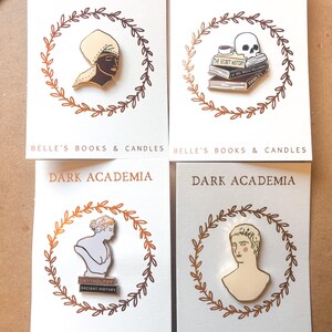 May include: Four enamel pins on white cards, each with a gold laurel wreath. Pins feature a bust, a skull on books, a figure with a head wrap, and a male bust. Cards read "BELLE'S BOOKS & CANDLES" and "DARK ACADEMIA".