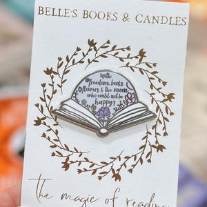 May include: A white card with gold text reading "BELLE'S BOOKS & CANDLES" and "the magic of reading." A pin in the shape of an open book is in the centre, surrounded by a floral wreath. The pin has the quote "With freedom books, flowers & the moon, who could not be happy?"