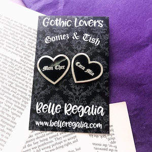 May include: Two heart-shaped black and silver pins with the phrases "Mon Cher" and "Cara Mia" on a black card with the text "Gothic Lovers Gomez & Tish" and "Belle Regalia". The background is a purple surface.