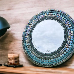 May include: A round mirror with a mosaic design in shades of blue, green, and iridescent colours. The mirror is surrounded by a border of small, colourful tiles.