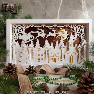 Puede incluir: Una escena navideña blanca en capas con una silueta de pueblo, el trineo de Papá Noel y una luna llena. El diseño está enmarcado en blanco con un fondo marrón. La parte inferior de la imagen tiene piñas, bellotas y una guirnalda de cuentas verdes.