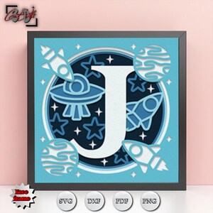 May include: A framed, layered paper art piece featuring a large white letter "J" surrounded by space-themed elements. The design includes rockets, planets, stars, and a UFO in shades of blue and white. The frame is black.
