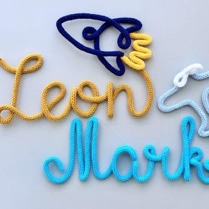 以下が含まれることがあります： 手編みの糸アート。筆記体で「Leon」と「Mark」の名前、青と黄色のロケット、水色の飛行機が描かれています。糸は黄色、青、白の色合いです。