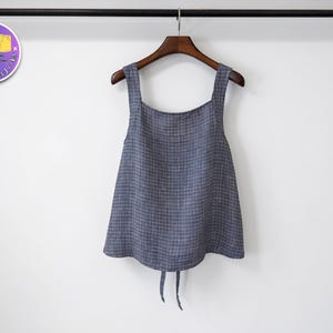 Könnte beinhalten: Ein blau-graues Tanktop mit Karomuster und breiten Trägern hängt an einem Holzbügel. Das Oberteil hat einen quadratischen Ausschnitt und eine ausgestellte Form. Der untere Teil des Oberteils hat zwei lange Bindebänder. Das Oberteil hängt an einer schwarzen Stange vor weißem Hintergrund.
