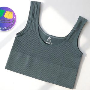 Puede incluir: Camiseta sin mangas acanalada de color verde azulado con cuello redondo y tirantes anchos. La parte superior es de color liso y parece estar hecha de un material elástico. Un logotipo circular con texto está en la esquina superior izquierda. La camiseta sin mangas es talla L.