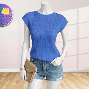 May include: A mannequin displays a blue textured top with denim shorts and a tan clutch. The top has short sleeves and a round neckline. The shorts are light blue denim. The clutch is rectangular. A logo is in the upper left corner.