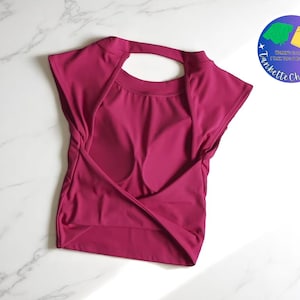 Puede incluir: Una blusa magenta con espalda abierta y mangas cortas. El frente tiene un diseño envolvente. Un logotipo circular con texto e ilustraciones está en la esquina superior derecha. La blusa probablemente es parte de una colección de ropa.
