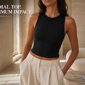 May include: A black sleeveless tank top with a crew neckline and a cropped hemline. The text MINIMAL TOP. MAXIMUM IMPACT. is visible in the upper left corner. The garment is paired with light beige pleated trousers with a high waist and side pockets.