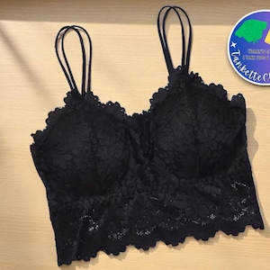 以下が含まれることがあります： 細いストラップとスカラップエッジが付いた黒いレースのブラレット。ブラレットは花柄で、明るい木材の表面に表示されています。右上隅にある円形のロゴには「Tankette Chic Looks」と「Women's Clothing Tank Tops & Bottoms」と書かれています。