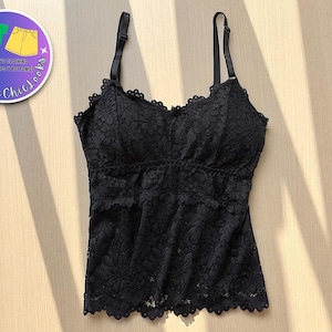 以下が含まれることがあります： 黒いレースのキャミソール、調節可能なストラップ付き。トップはスカラップのネックラインと裾が特徴です。この衣服は花のレース模様で作られています。画像には、「Tankette Chics」と「Women's Clothing Tank Tops & Bottoms！」と書かれたロゴも含まれています。