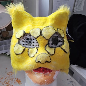 Puede incluir: Una máscara de gato amarilla hecha a mano con contornos negros y cubiertas oculares de malla. La máscara tiene una textura peluda y está diseñada para ser usada en la cabeza. El área de la boca es roja.