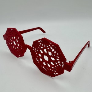 Puede incluir: Gafas de sol geométricas rojas con un diseño calado único. Las monturas y las patillas son de color rojo liso, y las lentes tienen un patrón distintivo. Las gafas de sol están sobre un fondo blanco.