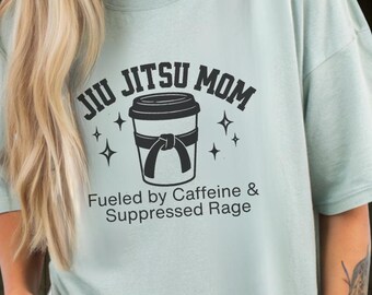 Può includere: T-shirt verde chiaro con la scritta "JIU JITSU MOM" sopra un disegno di una tazza di caffè con un nastro. Sotto, il testo recita "Fueled by Caffeine & Suppressed Rage".