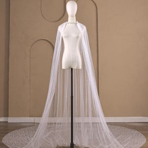May include: A floor-length white tulle cape featuring scattered pearl embellishments throughout the sheer fabric. The cape is secured at the neck with a decorative floral-inspired collar and drapes down to form a wide, circular train.