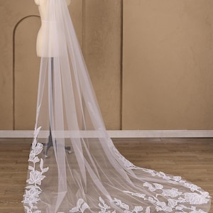 May include: A long, floor-length white tulle veil draped over a mannequin. The veil features intricate white floral embroidery along the edges and hem, creating a detailed pattern of flowers and leaves that extends across the bottom section of the fabric.